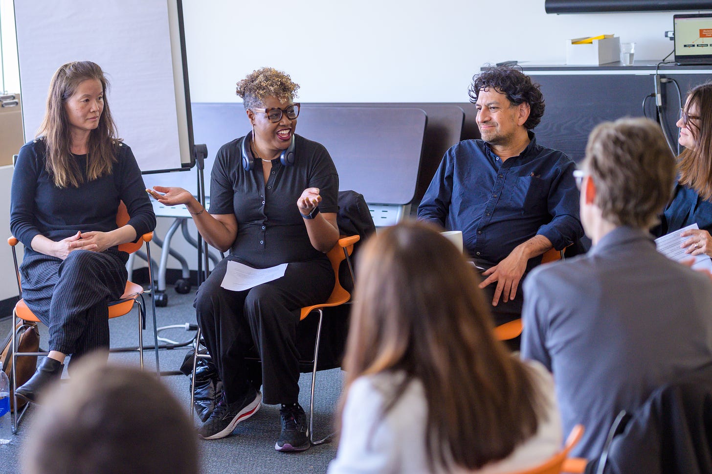 In a conference room, four people sit on orange chairs.