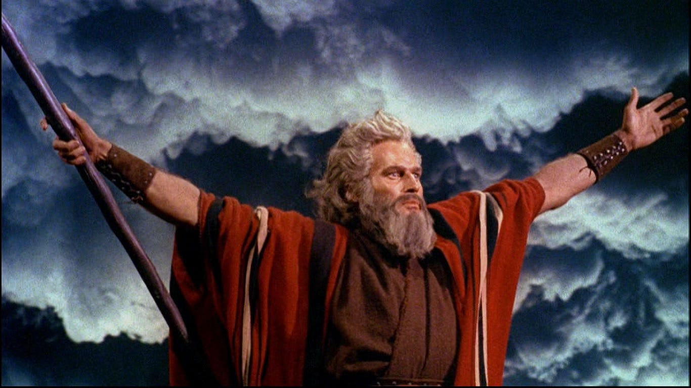 Color still photograph of Charlton Heston as Moses in The Ten Commandments with arms outstretched against a darkening sky, parting the Red Sea 