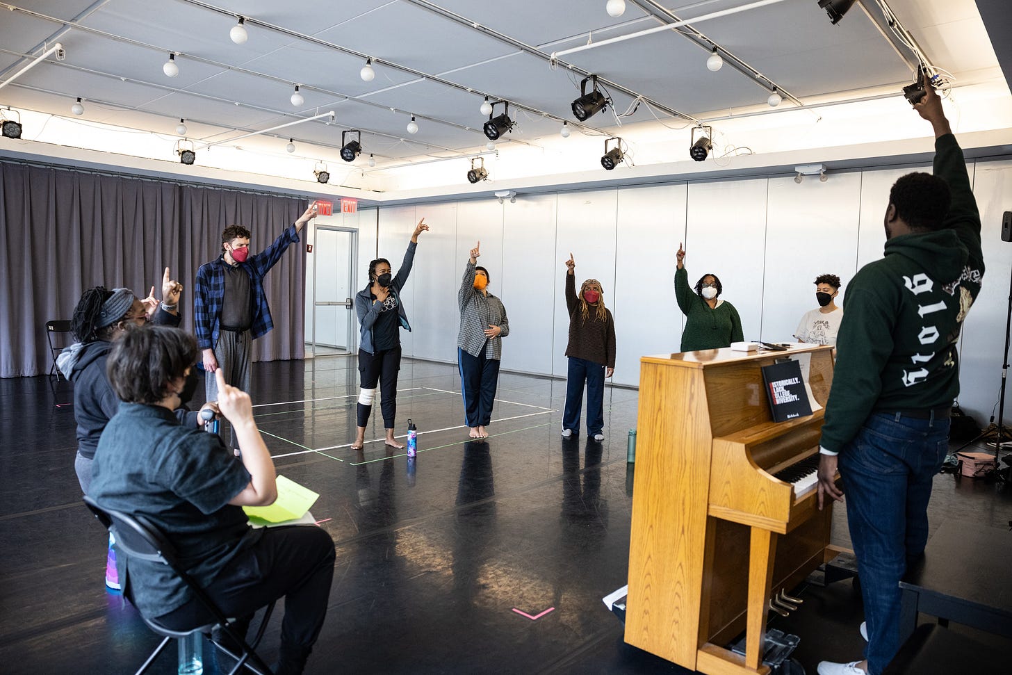 The cast of HBD/HPU are in a large circle around a piano; everyone extends a pointed finger to the sky as they rehearse vocals. Some performers stand, others sit in folding chairs, everyone is masked.