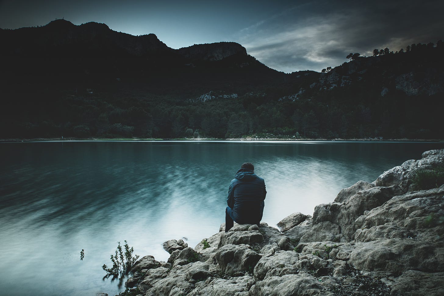 A man in a blue jacket sits on a rock ledge overlooking a lake. In the background are dark mountains.