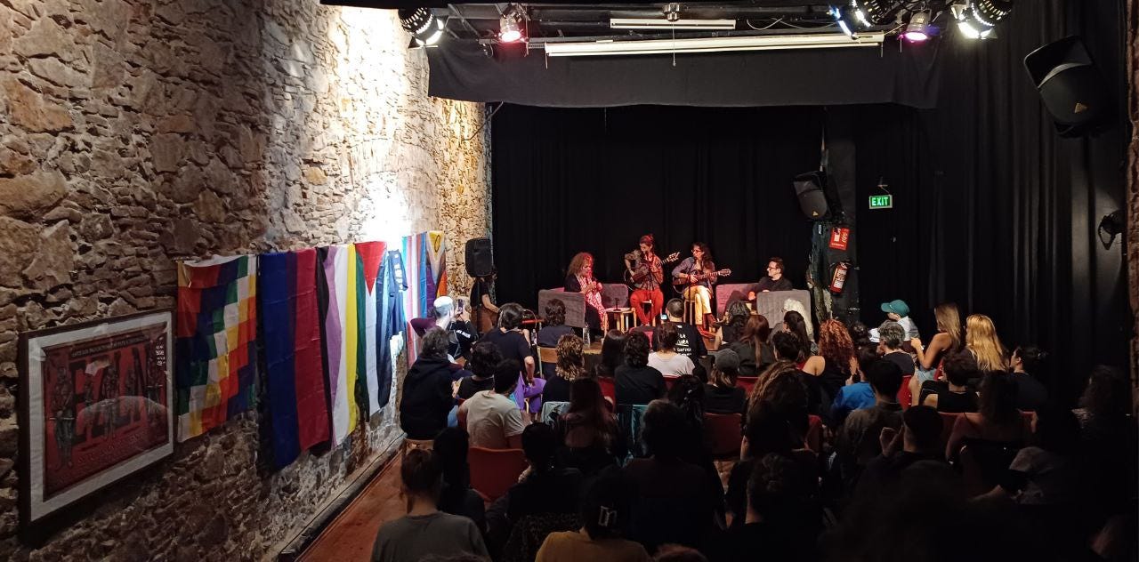 La sala piena di gente dell’Ateneu del Raval: sulla parete a sinistra diverse bandiere LGBTQ+ (bisessuale, trans, non binaria) e della Palestina; sul palco in fondo ci sono Susy Shock, il duo di chitarriste Bazán-Bonillo, e Brigitte Vasallo. 