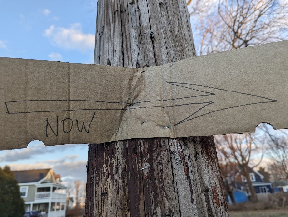 A piece of cardboard stapled to a pole. On the cardboard sign, someone has written the word "NOW" and drawn an arrow pointing to the right.