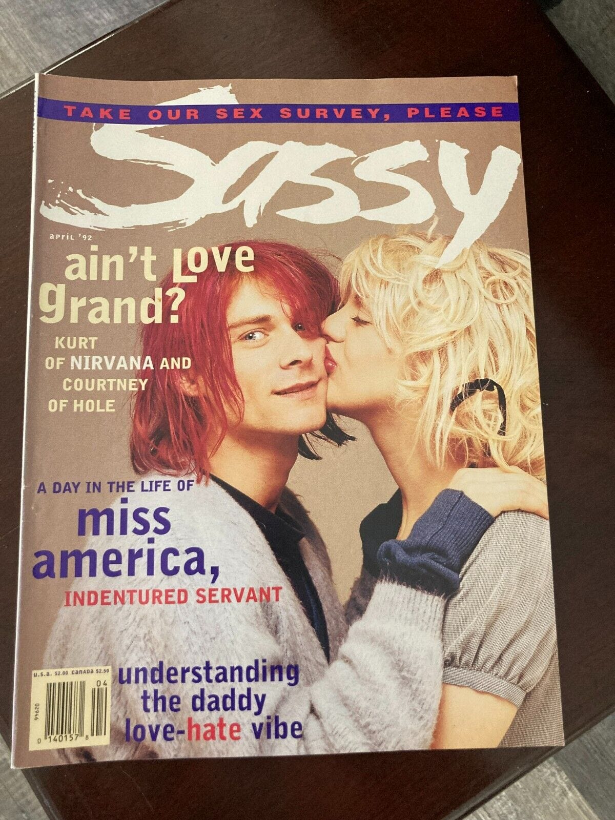 a copy of "sassy" magazine where kurt cobain, with messy dyed-red hair and a fuzzy cardigan, is kissed on the cheek by his wife, a blissed-out woman with tangled blonde hair and bright red lipstick