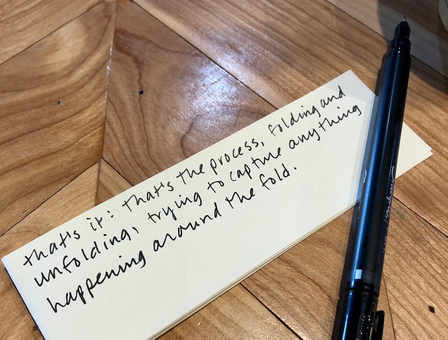 a few folded scraps of paper with a black pen set on their corner, all atop a wooden table. the final line of this newsletter is written on the paper in black ink.