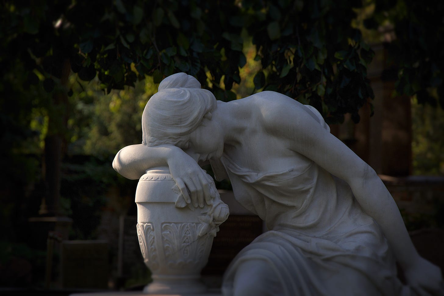 Close-up of marble statue in a park. A feminine figure is sitting. Her arm is braced on an ornate amphora next to her, and she’s resting her forehead on her arm. The pose suggests tiredness and sadness.