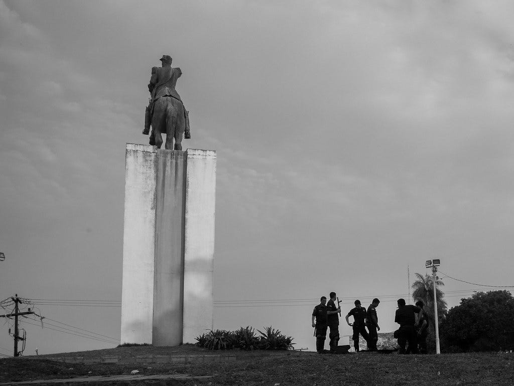 Black and white photo of a group of six police officers in uniform gathered under a plinth of a statue for a military officer Mariscal Lopez riding a horse in Asuncion, Paraguay. The officers are standing casually outlined against the sky and one can be seen carrying a baton, while another reaches for something in a back pocket.