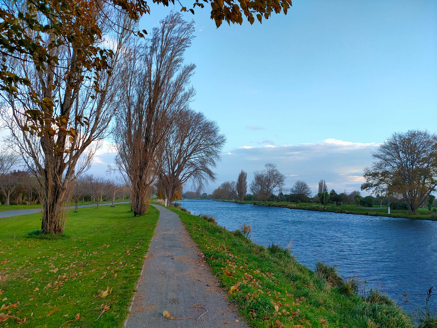 A path, a river, and green grass run parallel towards the distance. There are a few tall trees in the middle and far distance, all losing their leaves or bare. The sky is pale blue, and clouds are massing on the horizon.