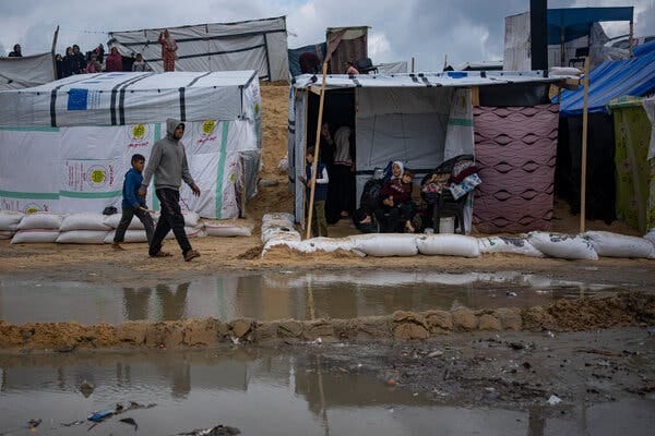 Several makeshift tents with people inside stand behind pools of dirty water.