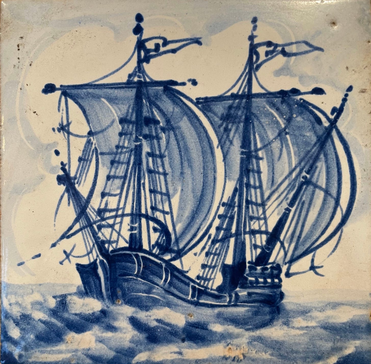 A blue and white tile of a tall ship with billowing sails and flags on the sea.