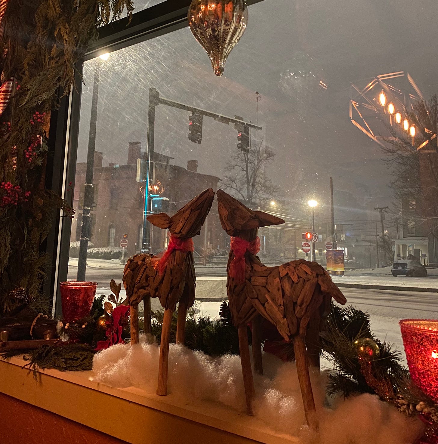 Christmas decorations, including two wooden reindeer touching noses, adorn a window. Out of the window, you can see a street corner covered in snow, and snow falling diagonally in the night. 