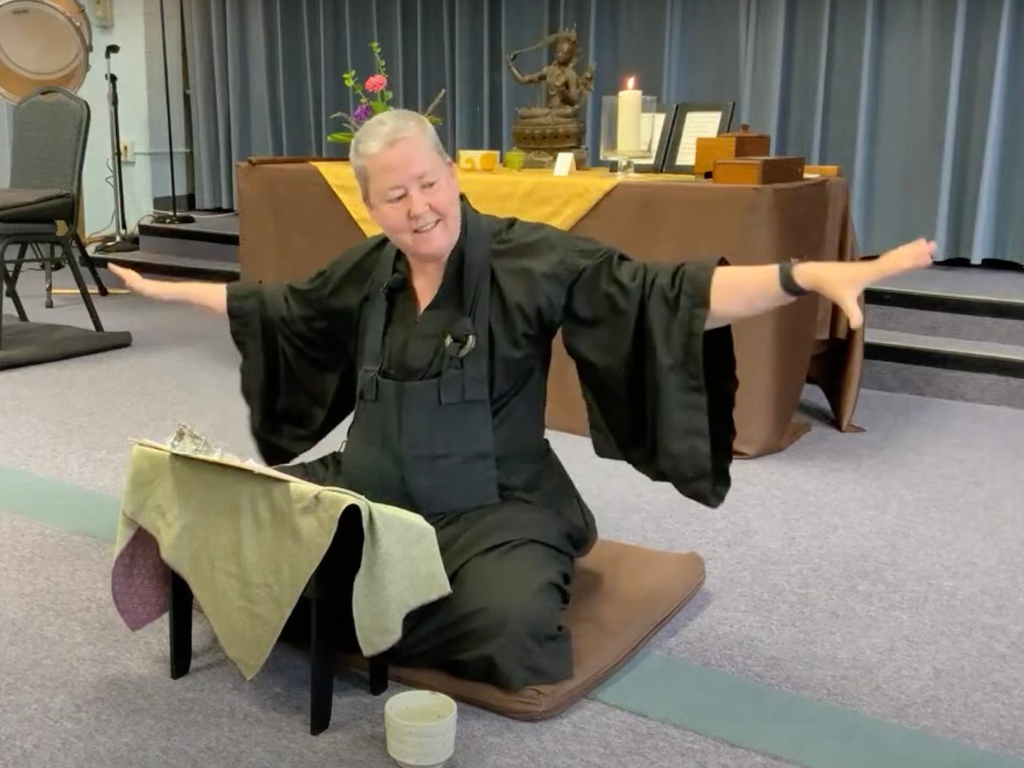 A video still of Sal Gessho Randolph giving a dharma talk. She is a white woman with short hair wearing black Zen sitting robes and a rakusu, smiling with her arms outstretched in a gesture as she speaks. She is seated on a cushion in a room with a purple-grey industrial carpet and behind her is a Zen altar with a bronze figure of Manjushi, a burning pillar candle, and flowers. 