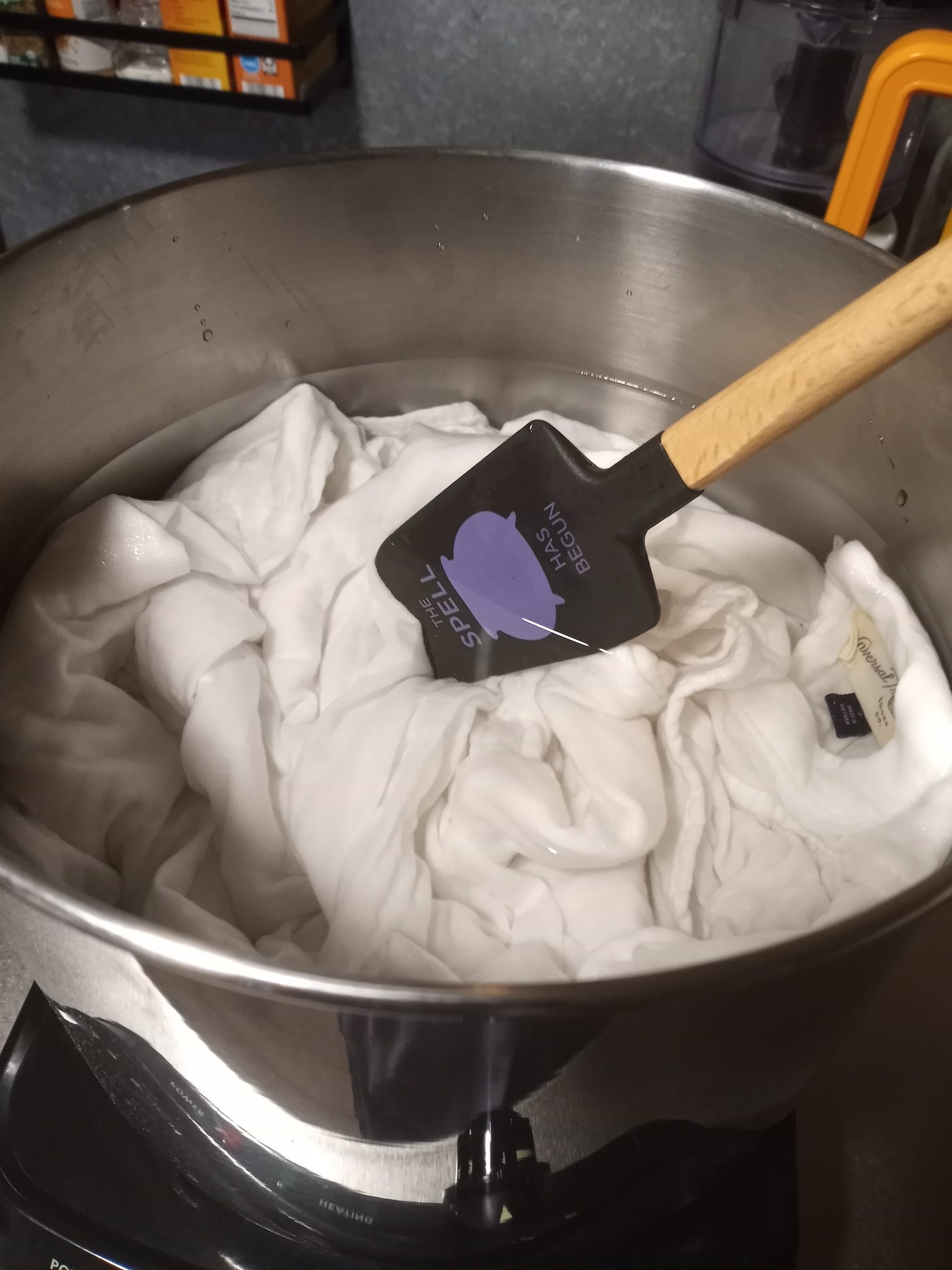 A white dress is submerged in water in a large metal pot, sitting on a burner. A spatula is stirring the mixture.