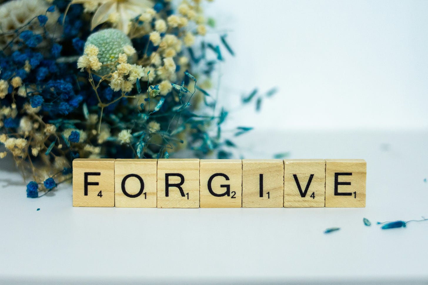 A pale background with blue, tan, and green dried flowers on the left with wooden Scrabble letters in the middle spelling out "FORGIVE"