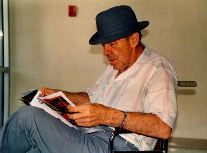 Older man reading a magazine, wearing a hat.