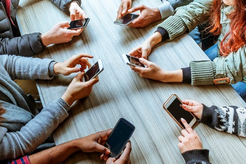 A group of people sit around a table. All you can see are their arms, with each holding a smart phone that is in use.