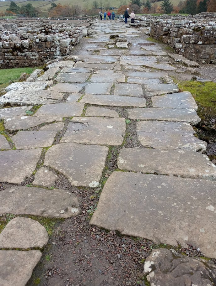 Paved road looking through remains of village, low walls of stone, toward tourists standing at what was once entrance to fort