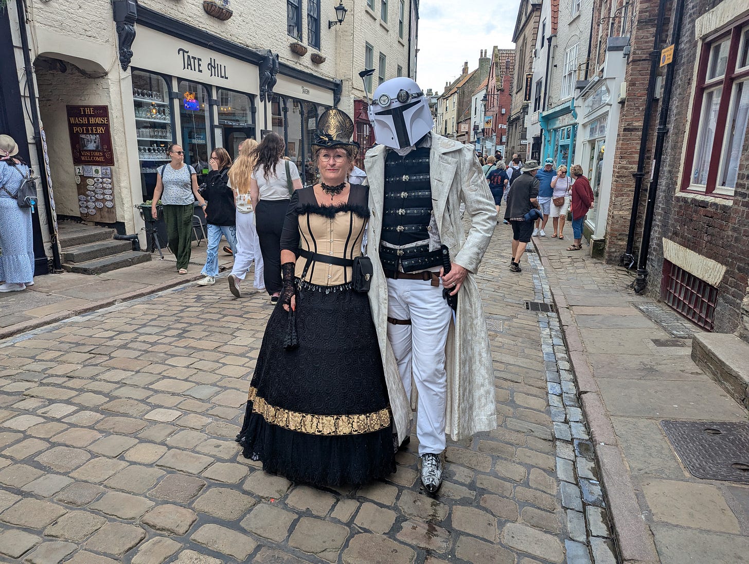 A steampunk couple pose for the camera on the street, one of them wearing a white Mandalorian helmet, modified slightly to look more steampunk