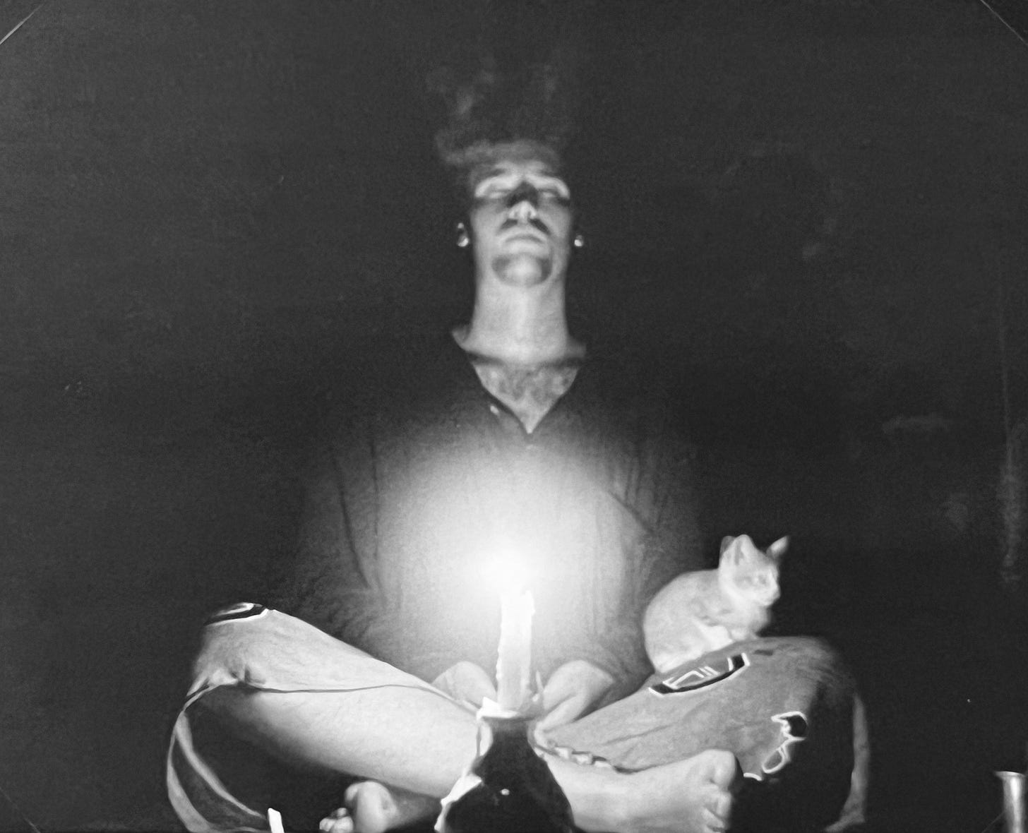 A black-and-white photograph of me sat meditating in a sarong in front of a candle stuck in the top of a bottle burning in the dark. On my left knee, a kitten is dozing within my hands are placed within my lap. The photo was taken in an off grid, meditation retreat close to the cliff edge on the southern side of Penang island.