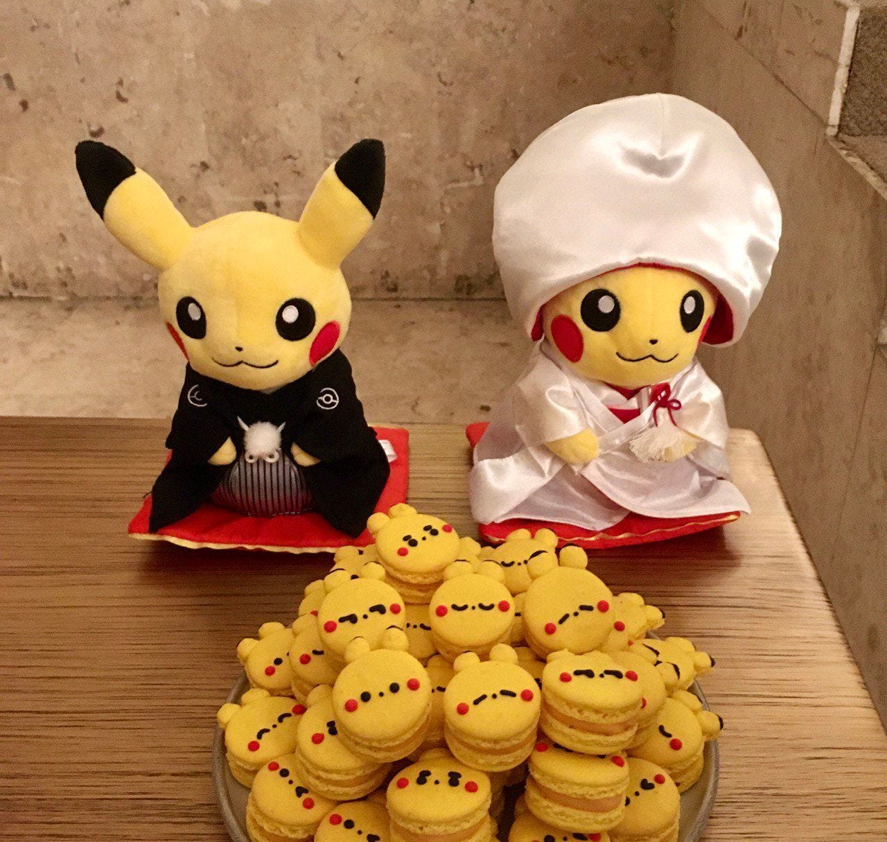 Foto de dois Pikachus de pelúcia com trajes tradicionais japoneses de casamento. Em frente a eles está um prato com macarrons amarelos decorados com carinhas do Pokemón.