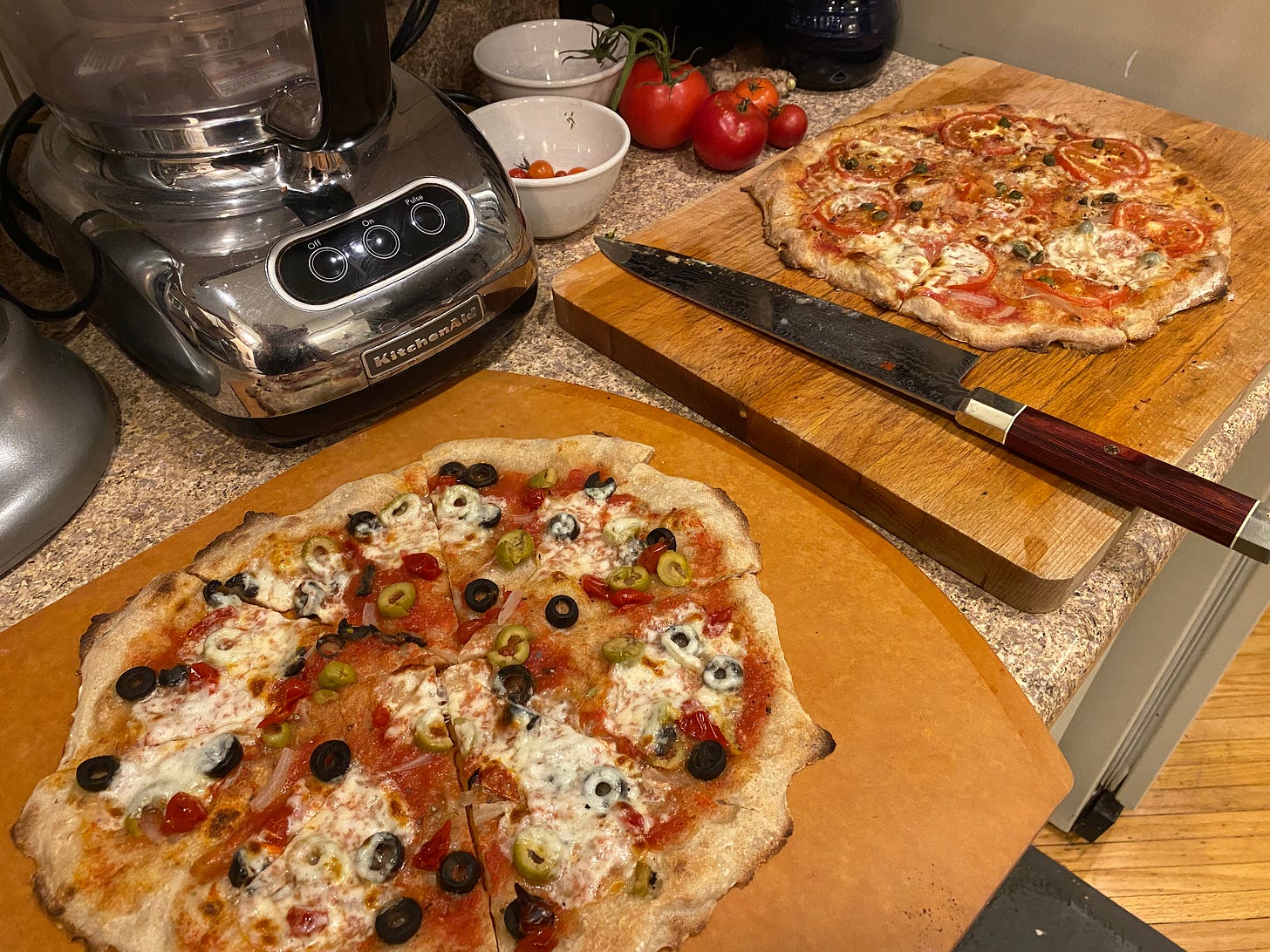 Two pizzas on boards with a knife in between them. In the foreground is the one with olives and peppers, and in the background is the one with tomatoes and capers, as described above. At the left of the frame are glimpses of a food processor and some fresh tomatoes on the vine.