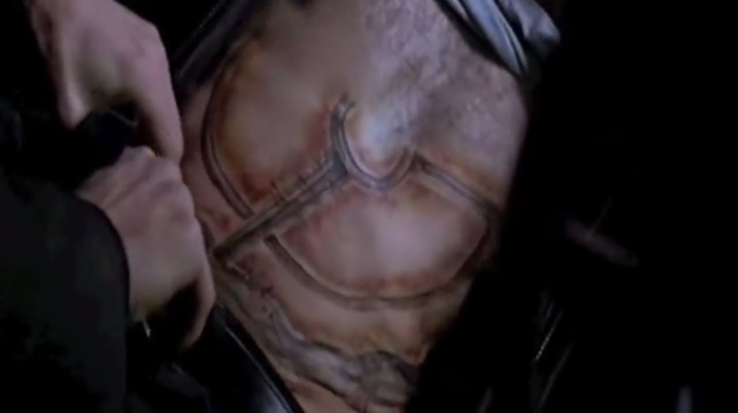Movie still from Crash. Close-up of a man's hands on another man's bare torso that has a bruised tattoo of a huge car emblem.