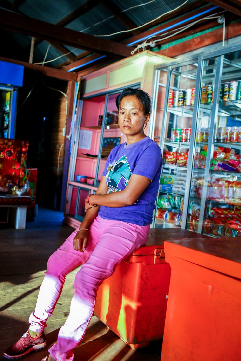 A woman in a purple t-shirt and pink pants looks extremely bored, sitting on a red cooler box in front of a glass cabinet filled with snacks. 