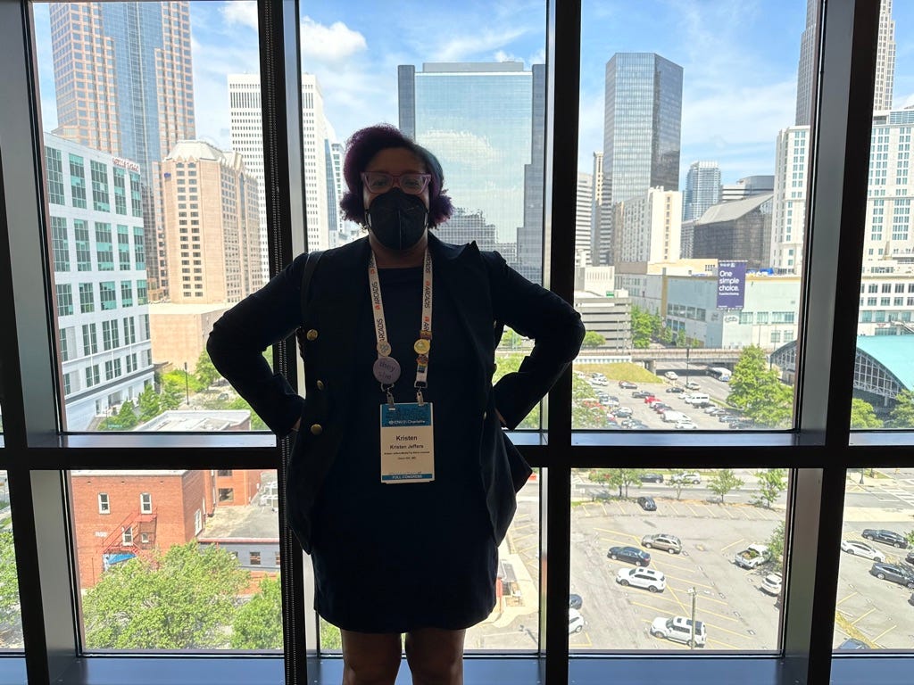 Kristen is standing in the Hyatt Place hotel lobby in front of a window with the Charlotte uptown skyline  behind her, with her hands on her hips. Her face is masked and she is wearing her CNU31 conference badge