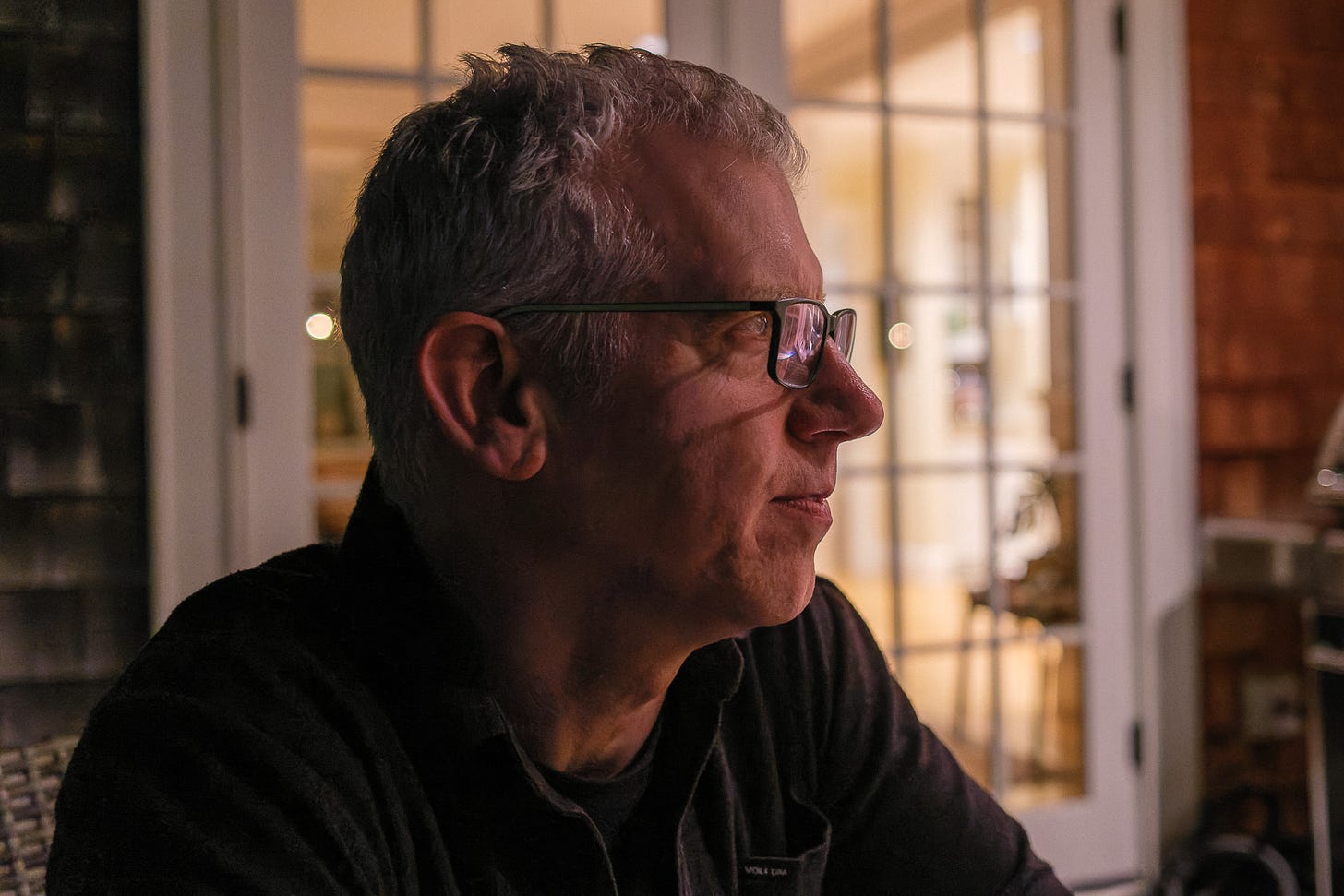 Profile photo of a man listening to a conversation late at night outside on a deck, head and shoulders framing, with the illuminated interior of a house in the background.