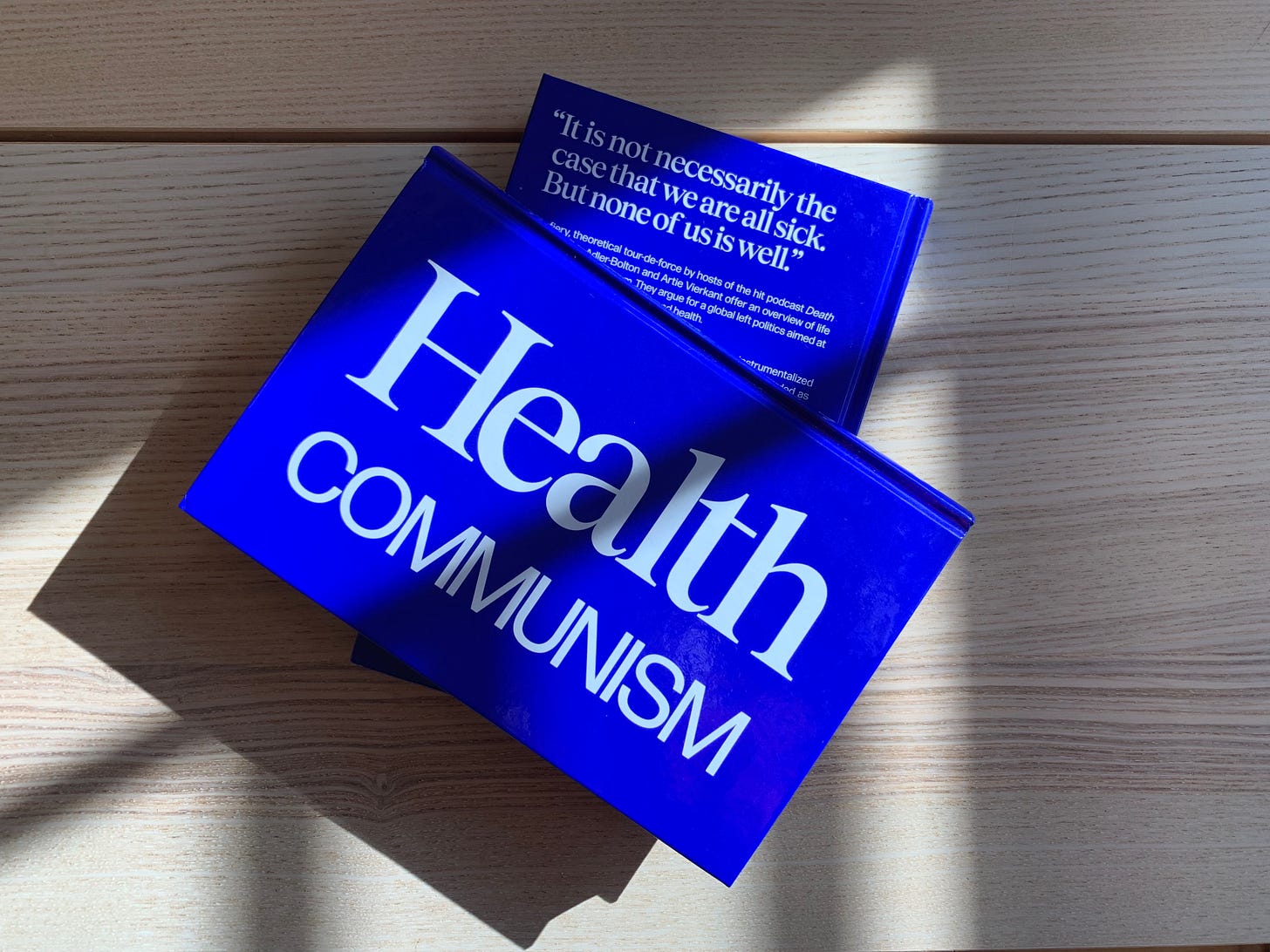 color photograph, two copies of Health Communism sit in sunlight and shadow on a light wood table. The book is bright blue with white lettering, the front cover, which is visible on the top copy, has a very simple design with the words Health Communism oriented along the spine instead of the top of the book. 
