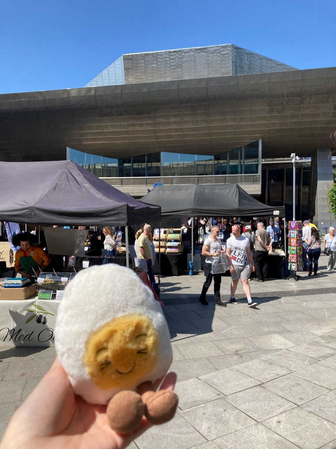 Photograph of a plush toy in the likeness of an egg with a smiley face in its yolk. In the background are a bunch of market stalls selling food and crafts outside an architectural theatre against a clear blue sky. It is busy and people are wearing sunglasses. 