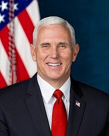 Official White House portrait. Pence is smiling in front of an American flag. He wears a black suit, red tie, and an American flag lapel pin.