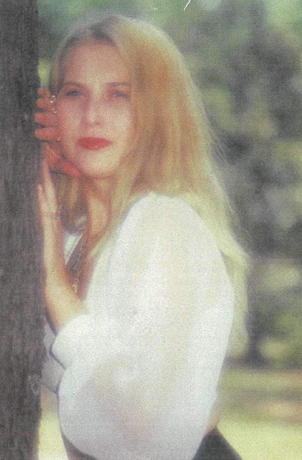 Photo of Tammy with long blonde hair and red lipstick posing against a tree in a white top