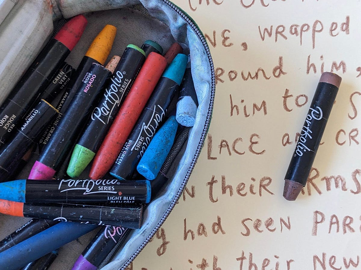 Photo of an open pencil case filled with colorful pastel crayons. It sits on an open sketchbook with brown writing on it.