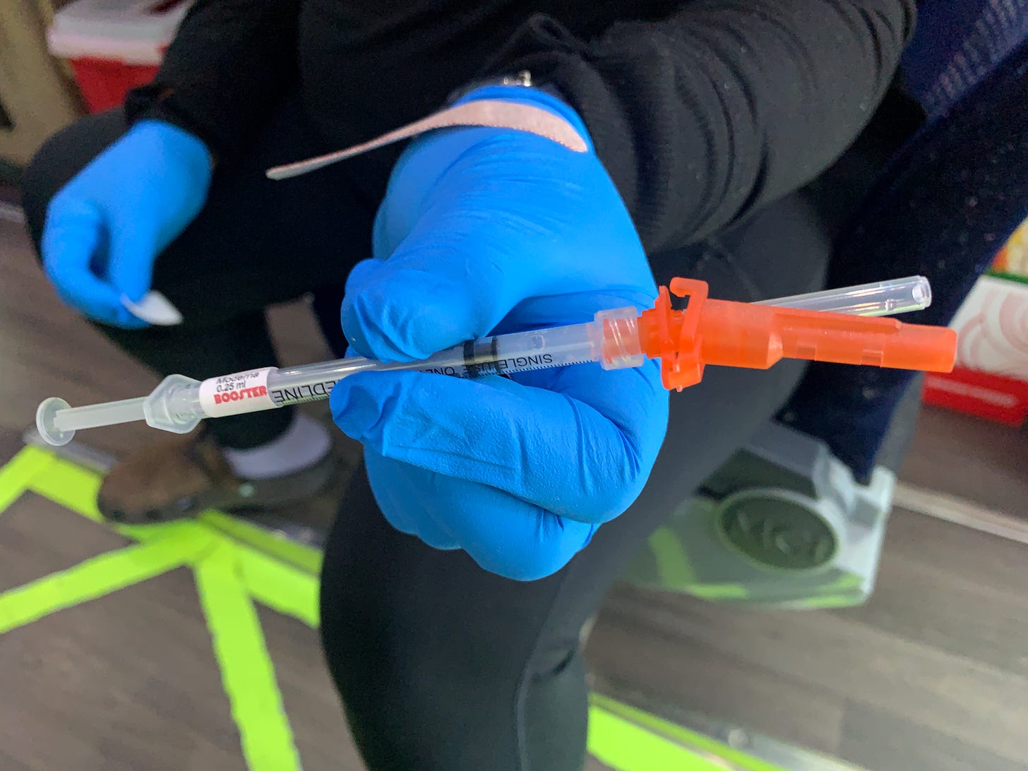 Syringe with a sticker saying "Booster" is held by a blue gloved hand. 