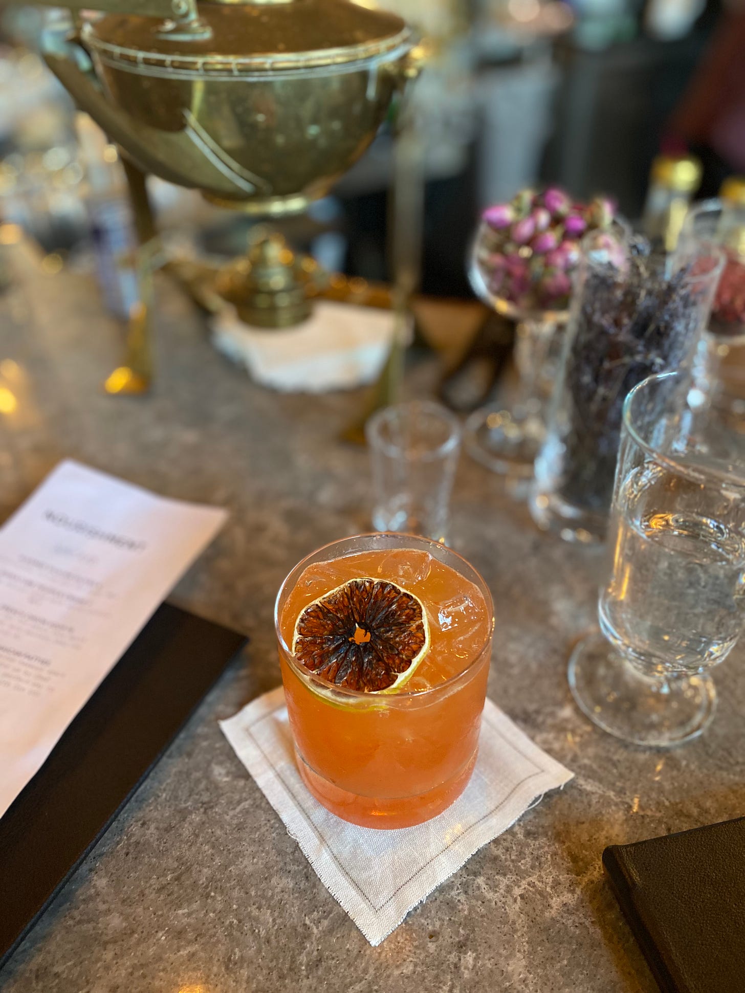 A cocktail with a dried orange slice in a highball glass, on a white napkin. There are other glasses nearby, including some filled with floral garnishes. On the left of the drink is a menu, and in the background is a small antique brass still.