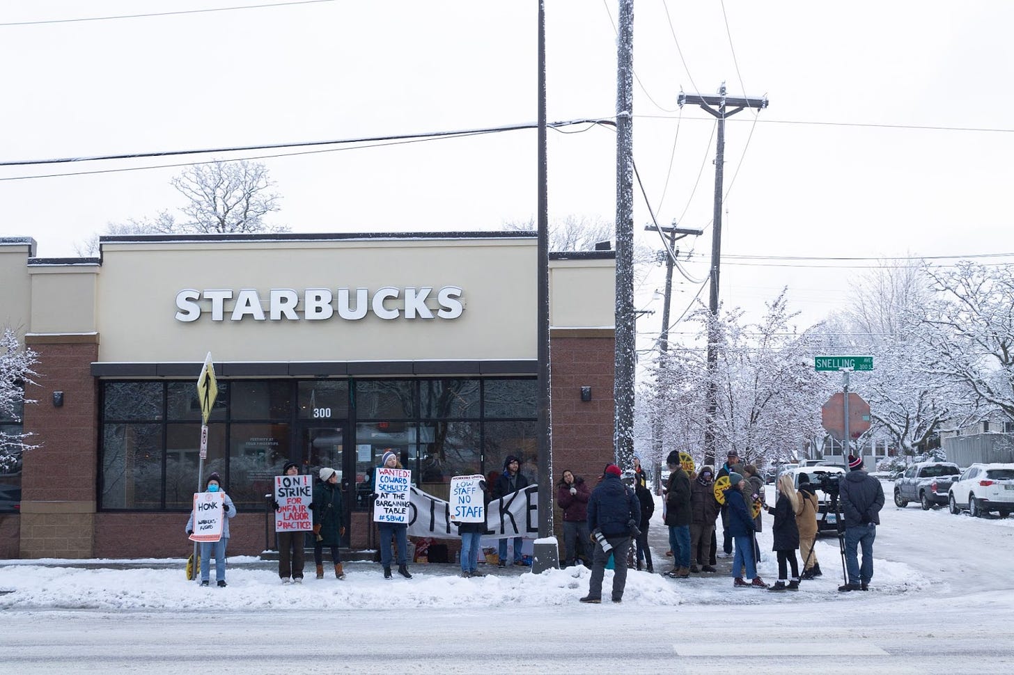 a small crowd pickets on the sidewalk in the snow in front of a starbucks location holding signs