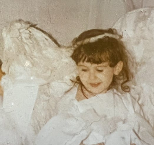 A faded photograph of young Lyric, in a white angel costume, made in protest by Lyric’s mother, after Lyric’s grandparents made Lyric a “devil” the halloween prior. Lyric loved the devil costume, despite Lyric’s mother’s objections. 