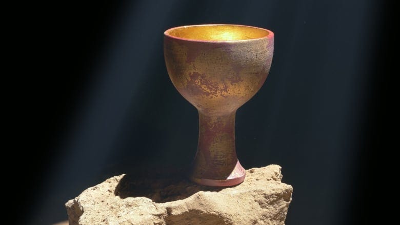 A metallic gold and pink-hued chalice stands empty on a stone, with rays of light shining down upon it. 
