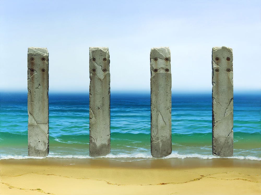 Four weathered pillars of concrete stand at ocean's edge. The sky is a hazy fog that blurs at the horizon above a deep blue sea. Approaching the foreground, sea green water becomes glassy reflecting the sun as the receding tide slowly rolls out depositing a meandering line of detritus on the wet sand.