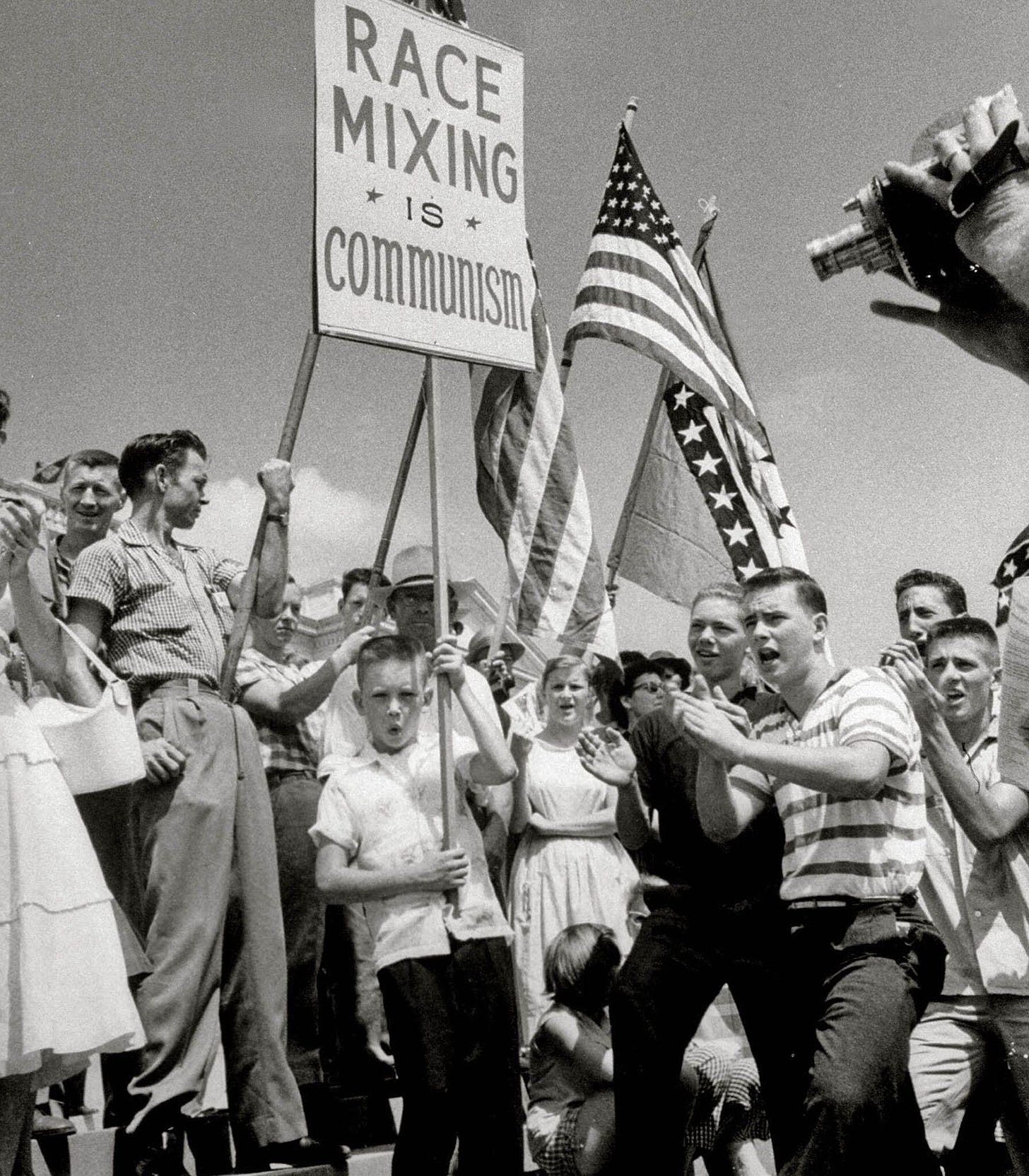 Black and white photo of a crowd of white people in the 1960s, one holding a sign that says “race mixing is communism” 