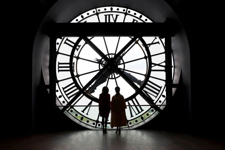 Two women stand silhouetted in front of a very large clock face.