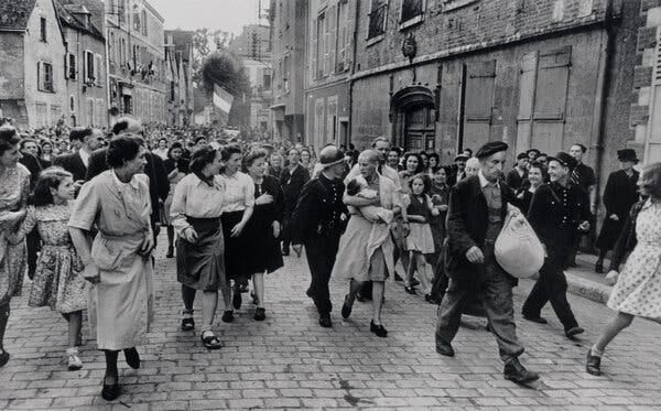 In a black-and-white photograph from 1944, a jeering crowd of people on a cobblestone street. At the center of the image, a woman with a shaved head cradles an infant.