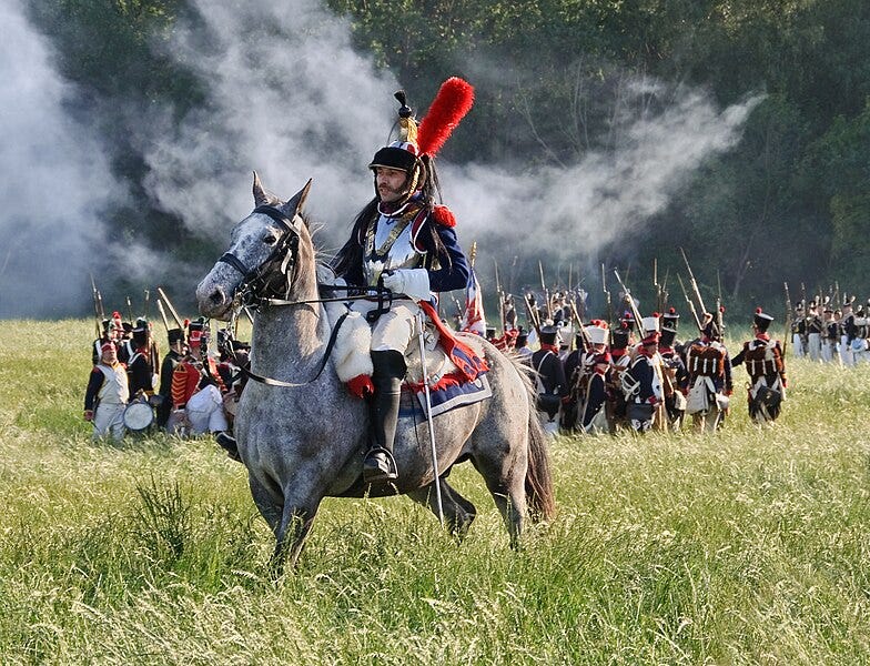 A man in colorful French military garb sits on a horse. Behind him are many men in similar uniforms carrying bayonets.
