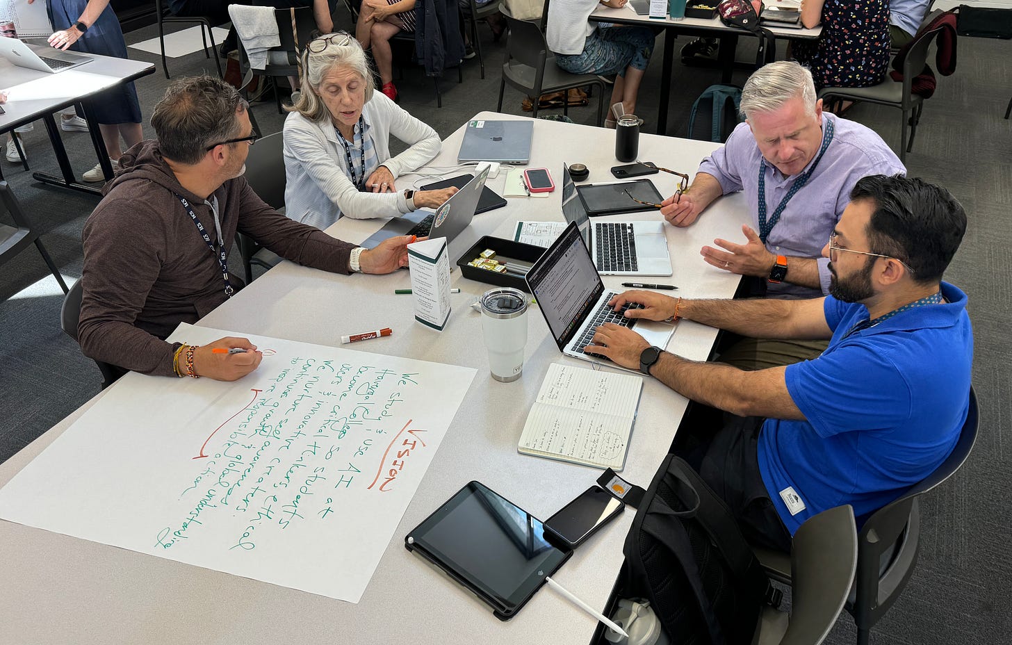 Four people are seated around a table, engaged in a discussion. The table has laptops, notebooks, markers, and a large sheet of paper with notes, including the word "VISION" in red and a list written in green marker. The group appears to be collaborating in a modern, well-lit workspace with other people and tables in the background.