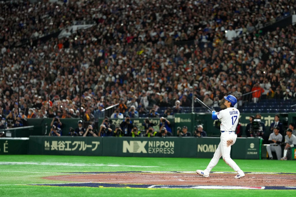 a photo of Shohei Ohtani with his bat out stretched. He is wearing a white Dodgers home uniform. Thousands of fans sit behind him and watch the ball carry over the fence for a home run.
