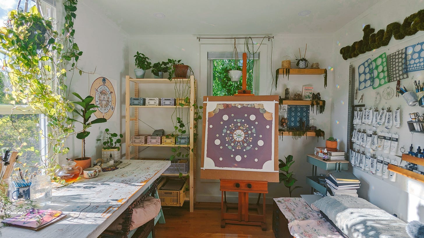 A shot of Caro's studio, with a long wooden work table and lots of paints lining the opposite wall. A painting in progress on a wooden easel, plants hanging near the windows. 