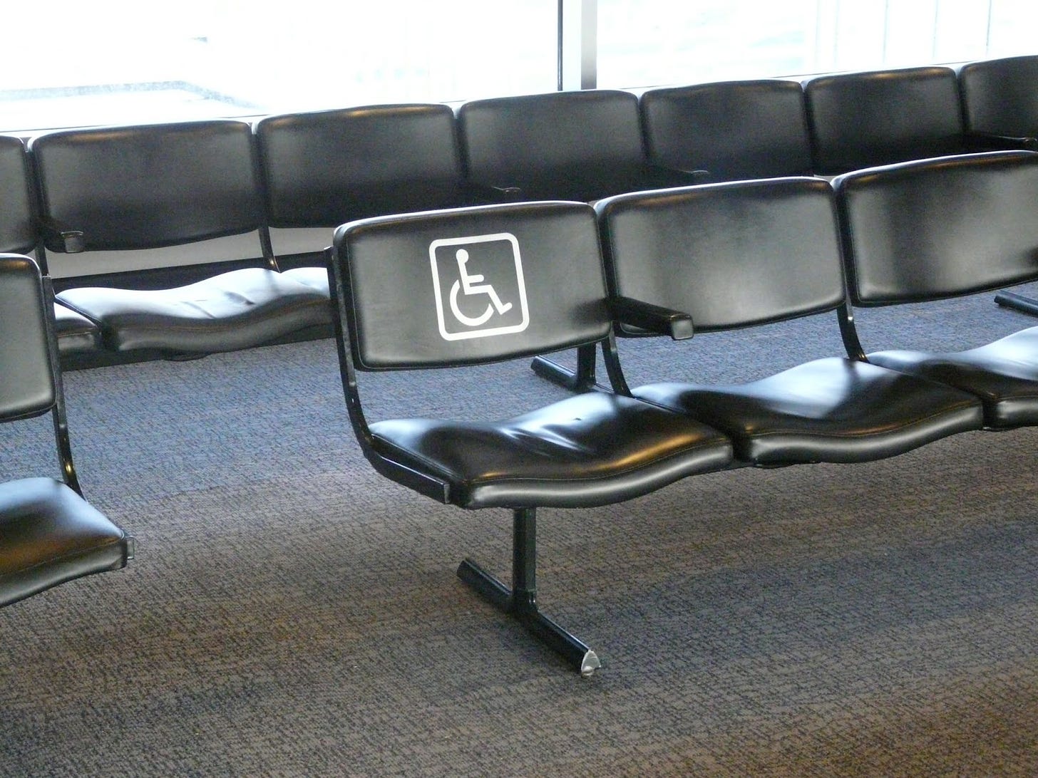 A photo that shows a row of black chairs at an airport. One chair has a pictogram on its backrest symbolizing a person in a wheelchair.