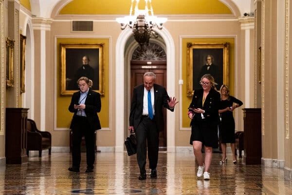 Senator Chuck Schumer wearing a suit and walking down a hall while gesturing. A person follows next to him.