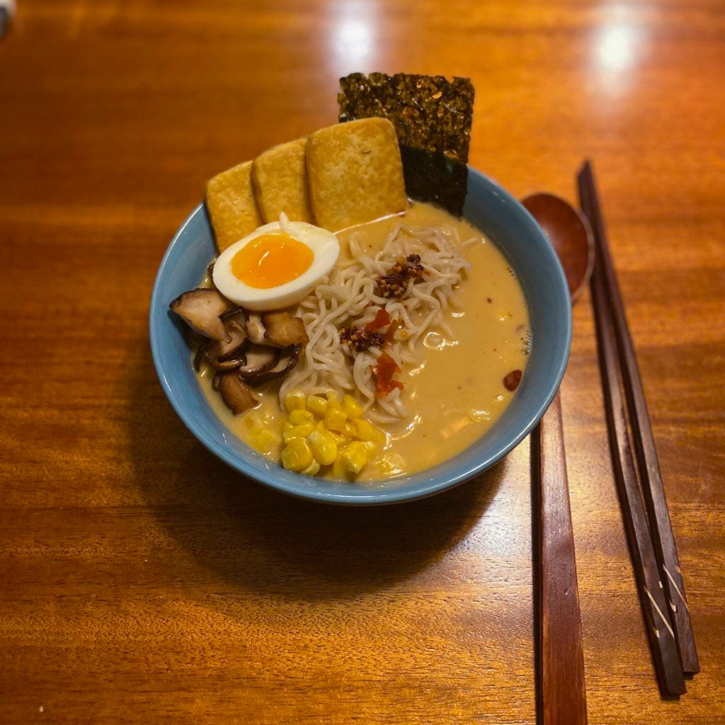 A blue bowl of ramen with a sheet of seaweed, slices of fried tofu, shiitake mushrooms, corn, and half a boiled egg arranged around the edges of the bowl. In the centre, on top of the noodles, is a small amount of chili crisp. Next to the bowl are dark wooden chopsticks and a matching spoon.