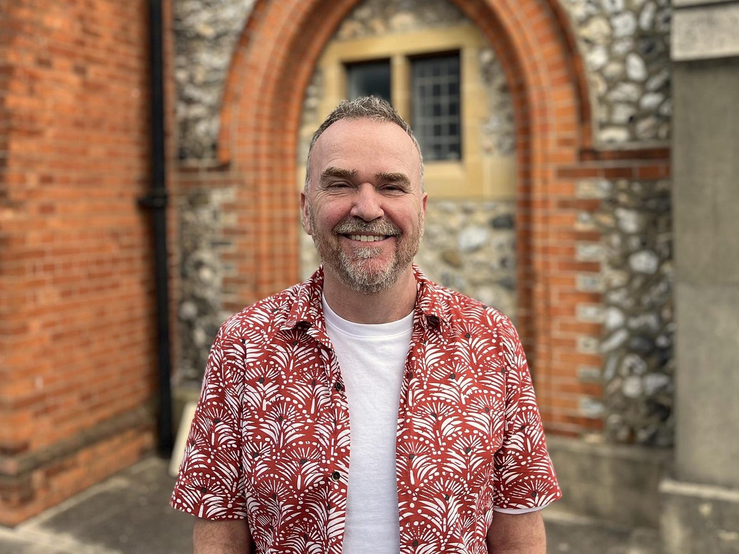 white man, middle aged, smiling outside church. Red highly patterend shirt.